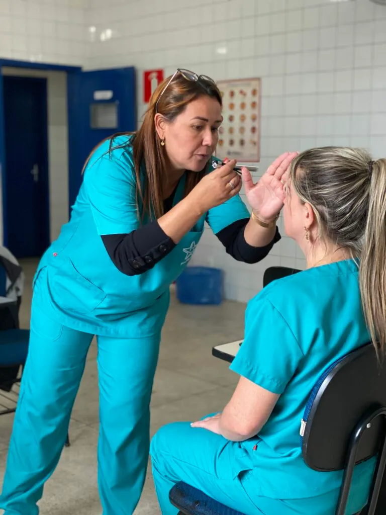 Mulher com uniforme médico azul-turquesa realiza um exame visual em uma cursista sentada.