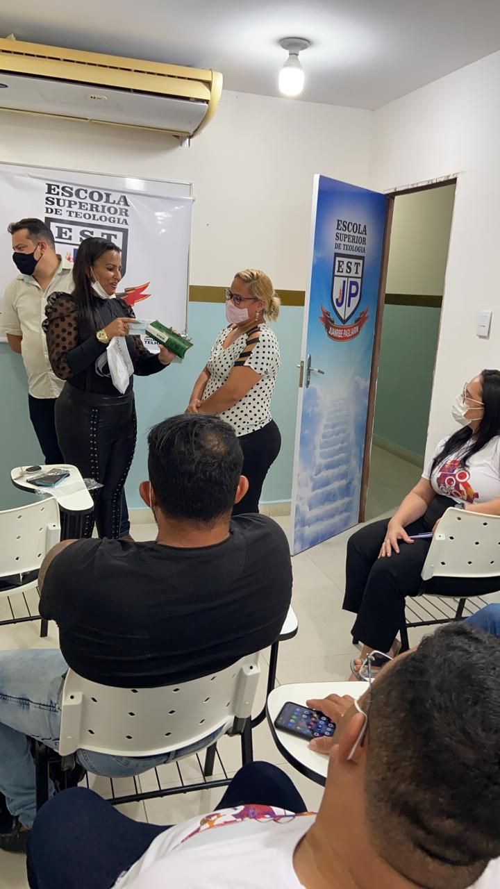 Uma mulher de blusa preta entrega um pacote a outra em uma sala de aula. Ao fundo, porta e banner trazem o logotipo 'Escola Superior de Teologia EST JP Juanribe Pagliarin'